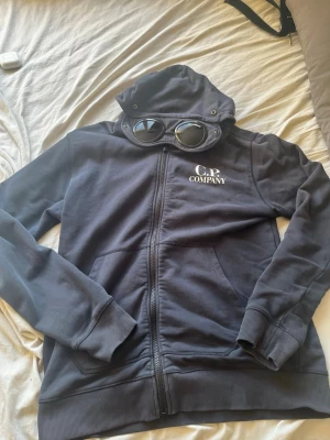 Mörkblå hoodie från C.P. Company - Säljer en mörkblå hoodie från C.P. Company med dragkedja och ikoniska goggles i huvan. Hoodien har ribbade muddar, fickor framtill och vit logga på bröstet. Perfekt för dig som vill sticka ut med en streetwear-look. 