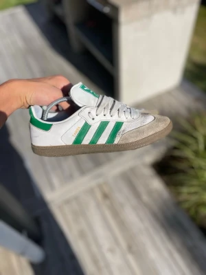 Adidas Samba sneakers  - Klassiska Adidas Samba sneakers i vitt med gröna detaljer och de ikoniska tre ränderna på sidan. Skorna har snörning, grå mockadetaljer på tån och gummisula. Perfekta för en sportig och retro look.