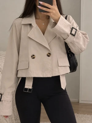 Kort beige trenchcoat med spännen - Trendig kort trenchcoat i beige med dubbelknäppning, stora knappar och dekorativa spännen vid ärmslut och midja. Klassisk krage och två fickor framtill. Perfekt för en stilren och modern look. Materialet ser ut att vara bomullsblandning.