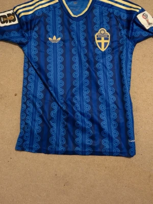 Blå Sverige fotbollströja - Säljer en blå  fotbollströja med Sveriges landslagsemblem på bröstet och klassiska gula detaljer. Tröjan har ett unikt mönster i olika blå nyanser, rund halsringning och korta ärmar. Perfekt för dig som vill sticka ut på matchen eller träningen. Pris kan diskuteras endast använd en gång nästan nytt skick 