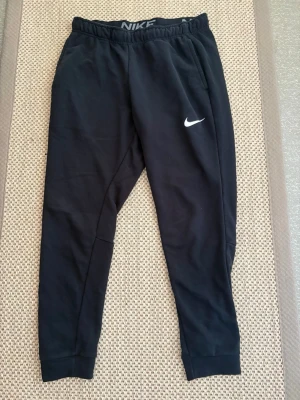 Svarta mjukisbyxor från Nike - Snygga svarta mjukisbyxor från Nike med vit logga på vänster ben. Byxorna har resår i midjan, smal passform och muddar vid bensluten. Perfekta för chill eller träning. Tillverkade i mjukt och bekvämt material. Bra skick 