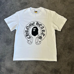Vit t-shirt med Chrome Hearts-tryck - Säljer en vit t-shirt med stort svart Chrome Hearts-tryck på bröstet och en ikonisk logga i mitten. T-shirten har rund hals och korta ärmar. Perfekt för dig som gillar streetwear och vill sticka ut med en unik design.