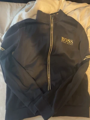 Hugoboss Kofta  - Snygg svart ziptröja från BOSS med gulddetaljer på dragkedjan och logga på bröstet. Tröjan har hög krage och ribbade muddar samt diskreta ränder på ärmarna. Perfekt för en stilren och sportig look.