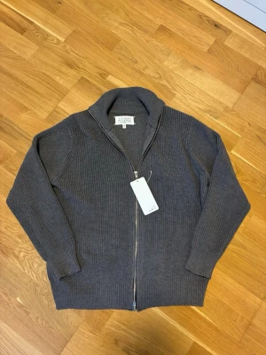 Maison Margiela zip tröja  - Säljer en stilren grå ziptröja från Maison Margiela. Tröjan är ribbstickad med hög krage och dragkedja framtill. Perfekt för lager på lager och har en clean look som passar till det mesta. Materialet känns mjukt och håller värmen bra.