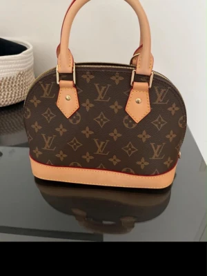  brun  handväska - Snygg handväska från  i klassiskt brunt monogrammönster med beigea detaljer och guldfärgad dragkedja. Väskan har rundad form, två handtag och detaljer i läder. Perfekt för dig som gillar lyxiga accessoarer och tidlös design.
