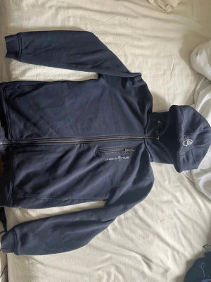 Marinblå hoodie från Sail Racing - Snygg marinblå hoodie från Sail Racing med dragkedja framtill och en extra ficka med zip på bröstet. Broderad logga på bröstet och huva med tryckt logga. Tillverkad i mjukt bomullsmaterial, perfekt för chill dagar.