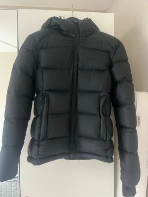 Svart pufferjacka med huva - Säljer en svart pufferjacka med huva och dragkedja framtill. Jackan är använd en vinter men max 10 gånger. Den är figursydd och väldigt varm. Nypris: 2500 kr. 