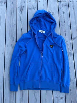 Blå hoodie Comme des Garçons Play - Blå hoodie med dragkedja från Comme des Garçons Play. Har en svart hjärtpatch med ögon på bröstet, två fickor fram och justerbar huva med snören. Tillverkad i mjukt material som känns skönt mot huden. Perfekt för en chill och stilren look.