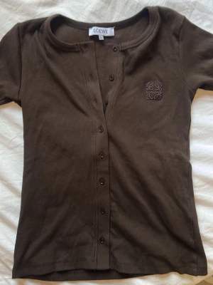 Brun ribbad T-shirt  topp från Loewe - Snygg brun ribbad topp från Loewe med korta ärmar och rund halsringning. Knäppning framtill med bruna knappar och Loewe-logga broderad på bröstet. Tillverkad i mjukt, stretchigt material som sitter skönt på kroppen.