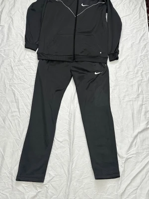 Nile tracksuit  - Säljer ett svart träningsset från Nike med zip hoodie och matchande byxor. Hoodien har dragkedja, fickor och vit Nike-logga samt vita detaljer över bröstet. Byxorna har rak passform och vit logga vid höften. Perfekt för träning eller chill.