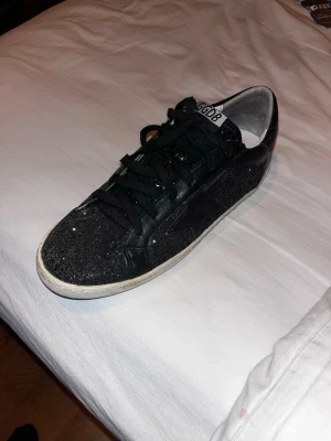 Svarta glittriga sneakers Golden Goose - Snygga svarta sneakers från Golden Goose med glittriga paneler och vita slitna sulor. Skorna har svarta skosnören, vit insida och detaljer i glitter på både sidor och häl. Perfekta för dig som vill sticka ut med en unik och trendig look. Väldigt unika skor i bra skick! Storlek 43 