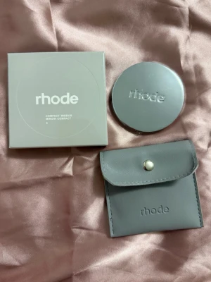 Rhode Compact Mirror med tillhörande fodral. - A stylish, minimalist compact mirror set from rhode, featuring a sleek grey design and branded details.