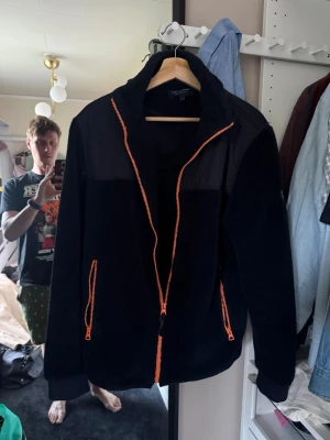 Svart fleecejacka från Lexington - Svart fleecejacka från Lexington med orange dragkedja och detaljer. Jackan har hög krage, två fickor med dragkedja och är tillverkad i mjukt, värmande fleecematerial. Perfekt för kyliga dagar och enkel att matcha med olika outfits.