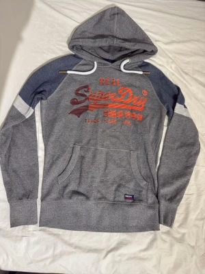 Superdry hoodie - Snygg grå hoodie från Superdry med kontrastmörkblå ärmar och vitt tryck på bröstet. Stor Superdry-logga i orange och rött framtill, samt känguruficka och vit dragsko i huvan. Perfekt för en avslappnad streetwear-look.