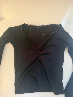 Svart långärmad topp från FB Sister - Svart långärmad topp från FB Sister med v-ringning och spetsdetaljer längs halsen. Toppen har en snygg rynkning framtill som ger en feminin touch. Perfekt att styla med jeans eller kjol för en trendig look.