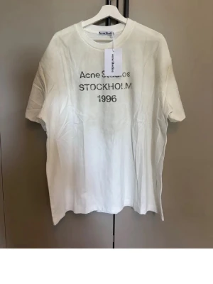 Vit Acne Studios t-shirt Stockholm - Vit t-shirt från Acne Studios med tryck 'STOCKHOLM 1996' på bröstet. Klassisk passform och rund halsringning. Tillverkad i 100% bomull, perfekt för en clean och stilren look.