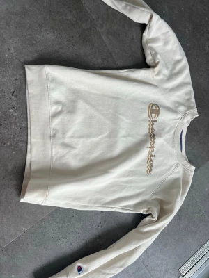 Vit sweatshirt från Champion - Säljer en stilren vit sweatshirt från Champion med broderad logga i beige och guld på bröstet samt liten logga på ärmen. Tröjan har rund halsringning, ribbade muddar och är gjord i mjukt bomullsmaterial. Perfekt för en avslappnad och sportig look.