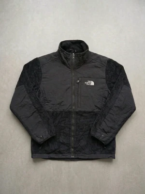 Svart teddyjacka från The North Face - Mysig svart teddyjacka från The North Face med hög krage och dragkedja framtill. Jackan har mjukt, fluffigt material på ärmar och rygg samt slätare tyg på bröst och axlar. Logotypen är broderad i vitt på både fram- och baksidan. Perfekt för kalla dagar.
