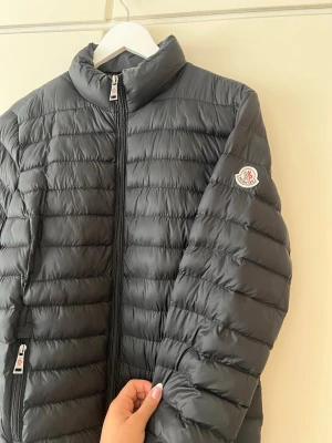 Svart dunjacka från Moncler - Snygg svart dunjacka från Moncler med quiltad design och hög krage. Jackan har dragkedja framtill, två sidofickor med dragkedja och Moncler-logga på ärmen. Perfekt för kalla vinterdagar och riktigt stilren look.