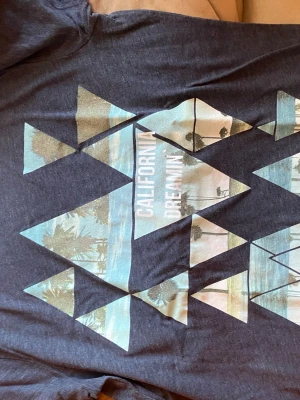 Mörkblå t-shirt med grafiskt tryck - Snygg mörkblå t-shirt med grafiskt tryck av trianglar och palmer samt texten 'California Dreamin'. Perfekt för dig som gillar avslappnad stil och coola motiv. Tillverkad i mjuk bomull för skön känsla hela dagen.