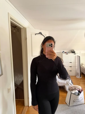 Svart ribbad polotröja - Säljer en svart, långärmad polotröja med ribbad struktur. Tröjan är figurnära och har hög hals, vilket ger en stilren och enkel look. Perfekt att matcha med jeans eller kjol för en clean outfit.