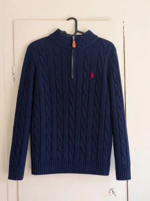 Stickad Ralph Lauren 1/4 zip - Säljer denna mörkblå kabelstickade tröja från Ralph Lauren med halvzip och den klassiska röda loggan. Tröjan är självklart 100 % äkta och har en tidlös och stilren passform som passar till det mesta. Skicket är överlag bra, men det finns några mindre skador/slitage! Därav säljs den till ett lägre pris.
