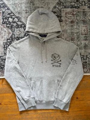 Grå hoodie från Polo Ralph Lauren - Snygg grå hoodie från Polo Ralph Lauren med huva och dragsnören. På bröstet finns ett tryck med en björn och texten 'POLO'. Hoodien har känguruficka och ribbade muddar. Perfekt för en avslappnad och trendig stil.