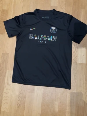 Svart PSG x Balmain t-shirt - Snygg svart t-shirt med PSG-logga och Balmain Paris-tryck i regnbågsskimrande färger på bröstet. Klassisk rund hals och korta ärmar. Perfekt för dig som gillar sportmode och streetwear.