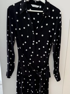 Skjortklänning medknytband - Klassisk och stilren svart midiklänning med vita prickar (polka dot) från & Other Stories (Paris Atelier). Modell i skjortstil med krage, knappknäppning framtill, långa ärmar med manschett och ett smickrande integrerat knytband i midjan som stängs fram eller bak. Perfekt för både vardag, jobb och finare tillfällen.