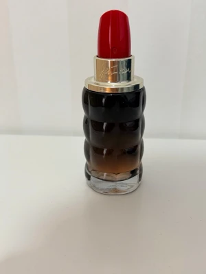 Cacharel Yes I Am Eau de Parfum 50ml  - Unik parfymflaska i glas med vågig svart till brun toning och ett lock som liknar ett rött läppstift. Flaskan har silverfärgad ring upptill och en modern, iögonfallande design som sticker ut på sminkbordet.