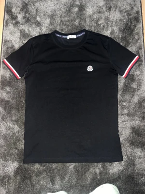 Svart Moncler t-shirt med logga - Svart t-shirt från Moncler med klassisk rund hals och korta ärmar. Ärmsluten har snygga rödvita ränder och bröstet pryds av Monclers ikoniska logga. Perfekt för en clean och stilren look.