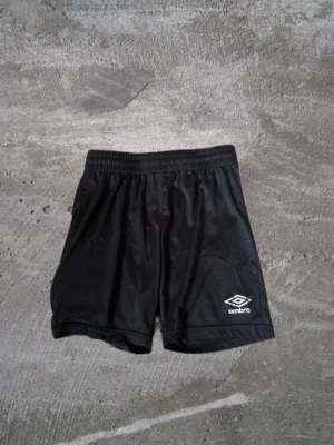 Svarta träningsshorts från Umbro - Säljer ett par svarta träningsshorts från Umbro med elastisk midja och klassisk logga på ena benet. Shortsen är tillverkade i ett lätt och glansigt syntetmaterial som passar perfekt för sport och aktivitet.