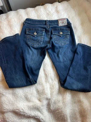 True Religion Blå bootcut jeans med fickdetaljer  - Snygga blå bootcut jeans med dekorativa bakfickor och kontrastsömmar. Jeansen har klassisk femficksdesign. Har sytt upp de sen innan därför är de lite kortare än originalet. Säljer för att de blivit för korta.💗