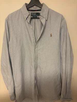 Randig skjorta från Polo Ralph Lauren - Klassisk blå- och vitrandig skjorta från Polo Ralph Lauren i slim fit. Skjortan har lång ärm, knappar framtill och broderad logga på bröstet. Tillverkad i bomull och passar perfekt till en clean och stilren look.