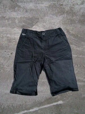 Svarta shorts med fickor - Säljer ett par svarta shorts med normal passform och klassisk design. Shortsen har två sidofickor fram och en bakficka, samt knapp och gylf framtill. Perfekta för sommardagar och enkla att matcha med olika stilar.