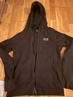 Svart hoodie från EA7 Emporio Armani - Svart hoodie med dragkedja från EA7 Emporio Armani. Hoodien har en huva och en liten gul EA7-logga på bröstet. Tillverkad i mjukt material som passar perfekt till en avslappnad stil.
