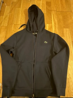 Svart hoodie med dragkedja Lacoste - Snygg svart hoodie från Lacoste med dragkedja framtill och klassisk krokodil-logga på bröstet. Tröjan har huva med dragsko och två fickor framtill. Tillverkad i mjukt och bekvämt material, perfekt för en avslappnad stil.