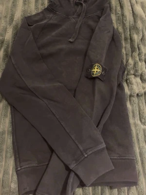 Svart hoodie från Stone Island - Säljer en svart hoodie från Stone Island med klassisk huva och dragsko. Hoodien har en enkel och clean design utan synliga tryck eller mönster. Perfekt för dig som gillar streetwear och vill ha något stilrent. Materialet känns mjukt och bekvämt.