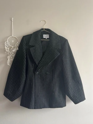 Randig kavaj/kappa - Randig kort kappa med markerad midja, oversized, något vadderade axlar, fickor i sidsömmen, ett innerfoder och en dubbel bröstknäppning.   Yttertyg: 91% polyester (60% Recycled polyester GRS), 8% akryl, 1% ull. Går att använda som en ”kavaj” men även som kappa utomhus