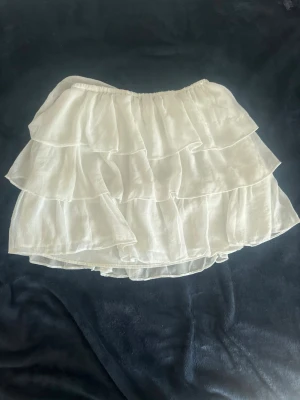 Vit volangkjol från Gina Tricot - Supersöt vit kjol från Gina Tricot med flera lager volanger och elastisk midja. Kjolen är kort och har en luftig känsla, perfekt för varma dagar. Tillverkad i ett lätt tyg som ger ett flowy intryck.