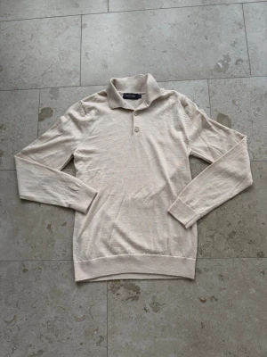 Beige stickad tröja från Dressmann - Snygg beige stickad tröja från Dressmann i mjuk merinoull. Tröjan har lång ärm, klassisk krage och två knappar framtill. Perfekt för dig som gillar stilrena och enkla plagg med en tidlös look.