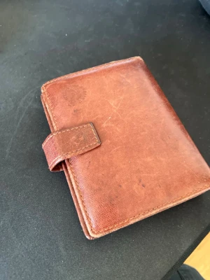 Plånbok äkta älgskinn - Säljer en klassisk brun plånbok i äkta läder med tryckknappsstängning. Plånboken har flera kortfack, plastfickor för ID och sedelfack. Insidan har ett tygfoder i grönt med mönster. Perfekt för dig som vill ha en stilren och praktisk plånbok.