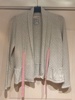 Ljusgrå kofta med rosa knytband - Ljusgrå kofta med diskret mönster, långa ärmar och volangkant nedtill. Rosa knytband framtill och en söt broderad kattpatch i nacken. Perfekt för dig som gillar gulliga detaljer och mjuka material.