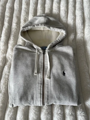 Ljusgrå zip hoodie från Polo Ralph Lauren - Tröjan har några fläckar som kanske går bort i tvätten men stan inte på avstånd.Snygg ljusgrå hoodie med dragkedja från Polo Ralph Lauren. Klassisk design med huva och dragsnören, två fickor framtill och den ikoniska broderade loggan på bröstet. Tillverkad i mjukt bomullsmaterial för maximal komfort. Tröjan är i storlek L men passar M