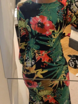 Lång blommig klänning med öppen rygg - Färgstark långklänning med blommigt mönster i grönt, rött, gult och vitt på mörk botten. Klänningen har lång ärm. Har öppen rygg
