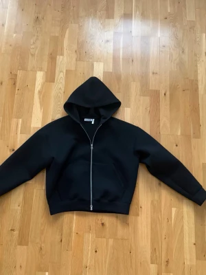 Weekday Scuba Hoodie - M - Stilren svart hoodie med dragkedja framtill och stor huva. Hoodien har en rymlig magficka och breda muddar vid ärmslut och nederkant. Materialet ser tjockt och mjukt ut, perfekt för en avslappnad look.