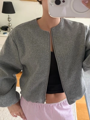 Grå bomberjacka  - Snygg grå bomberjacka med rund halsringning och dragkedja framtill. Jackan har en loose passform och långa ärmar, perfekt att slänga på över en t-shirt eller hoodie för en clean look.