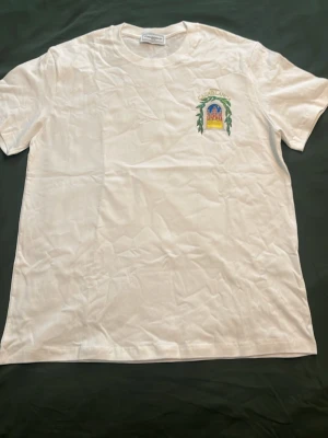 Vit Casablanca T-shirt - Säljer en vit t-shirt med färgglatt tryck av en byggnad och texten 'Avenida Casablanca' på ryggen. Trycket syns även i mindre format på bröstet. T-shirten har rund hals och korta ärmar, perfekt för dig som gillar unika motiv.