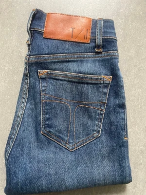 Blå jeans modell Kelly 24/32 - Snygga blå jeans i modellen Kelly med klassisk femficksdesign och kontrastsömmar. Jeansen har en mörkblå tvätt och en patch i brunt läder baktill i midjan. Perfekta för dig som gillar en tidlös och enkel stil.