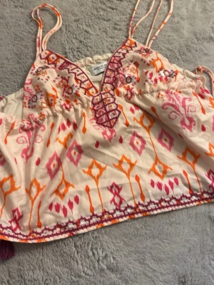 Broderad linnetopp med snörning - Säljer en färgglad linnetopp med broderade detaljer i orange, rosa och lila. Toppen har tunna axelband, v-ringning med snörning framtill och tofsar i lila. Mönstret är boho-inspirerat och materialet känns lätt och somrigt.